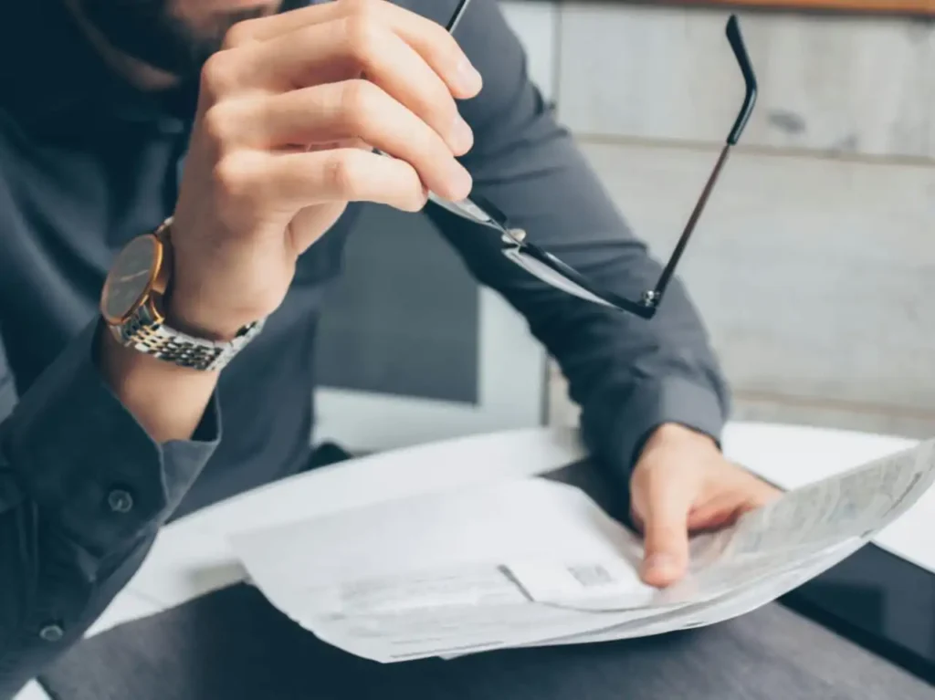 A person in a grey dress shirt holding glasses and reviewing paperwork at a desk.