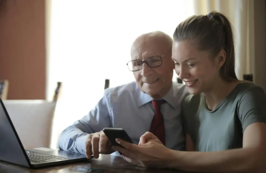 A young woman smiling as she shows an elderly man in a shirt and tie how to use a smartphone, with a laptop nearby on the table.
